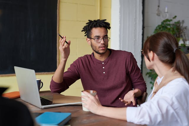 two people sitting at a table one person sitting in front on a laptop with a pen in their hand